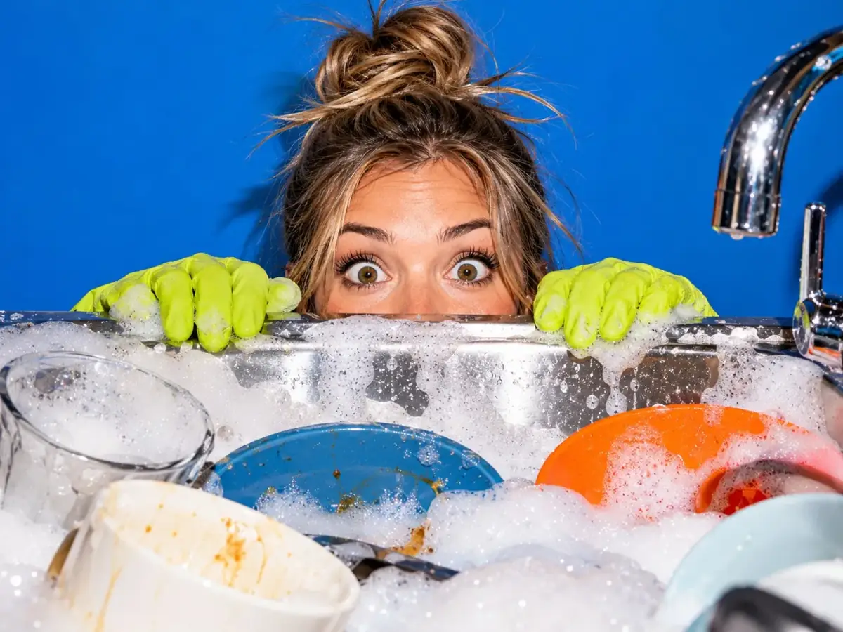 Woman in lime rubber gloves peeking over a sink full of soap suds and dishes